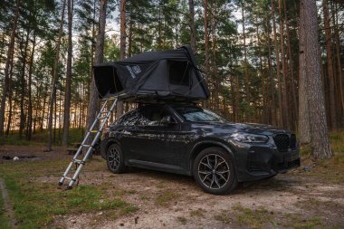 Ormana park edilmiş siyah bir BMW X4 2025. Çadır ve merdiveni de var. Uzun çam ağaçları ve sıcak ışık açık havada huzurlu bir ortam yaratır..