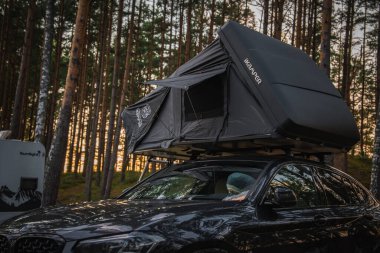 Siyah bir BMW X4 2025 IKAMPER çatısı olan bir çadır ormanlık alana park edilmiş. Sıcak bir günbatımında uzun çam ağaçları ve bir karavan görülebilir..