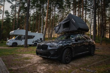 Çatısı çadırı olan siyah bir BMW X4 2025 çam ormanlarının kamp alanına park edilmiştir. Beyaz bir karavan ve yumuşak güneş ışığı doğal ortamı güçlendirir..