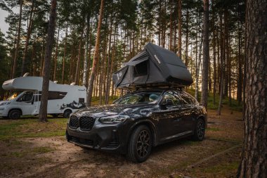 Siyah bir BMW X4 2025 Çam ormanı kamp alanında park etmiş bir çatı üstü çadırı. Arka planda sıcak ışık altında beyaz bir karavan görünüyor..