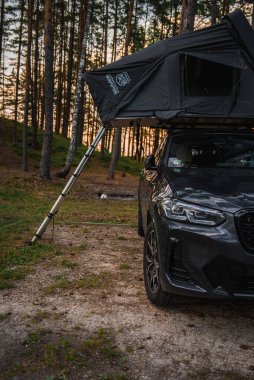 Siyah bir BMW X4 2025 ormanlık bir alana park edilmiş ve çatısı merdiven destekli bir çadır. Gün batımı ışığı uzun çam ağaçlarının arasından çakıl zemine süzülür..