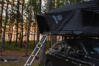 Siyah bir BMW X4 2025 iKamper çatısı çadırı olan bir orman. Güneş ışığı uzun çam ağaçlarının arasından süzülüyor..