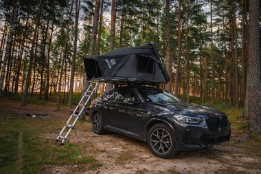 Siyah bir BMW X4 2025. Çatısı çadır ve merdiveni tenha bir ormana park edilmiş. Aracı çevreleyen uzun çam ağaçları sıcak gün doğumu ya da gün batımı ışığı altında.