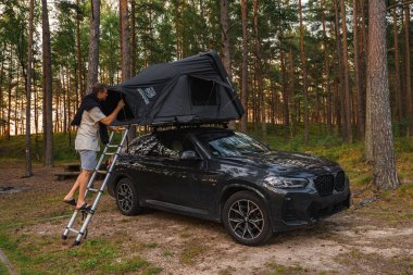 Siyah bir BMW X4 2025, yüksek çam ağaçları olan ağaçlık bir alana park edilmiş, çatısı olan bir çadır ve sıcak gün batımı ışığı altında merdivene tırmanan bir adam..