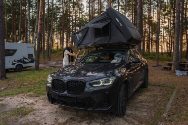 Siyah bir BMW X4 2025 IKAMPER çatısı olan bir çam ormanına park edilmiş bir çadır. Bir adam karavan ve kamp malzemelerinin yanındaki çadırla etkileşime geçiyor..