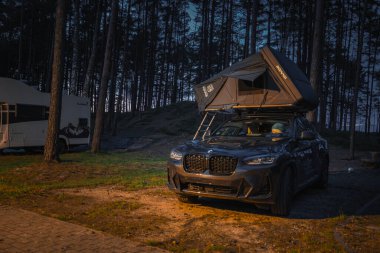Bir BMW X4 2025. Ormandaki asfalt bir alanda açık bir çatı üstü çadırı var. Arka planda uzun çam ağaçları ve bir karavan kamp alanını güçlendiriyor..