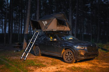 Çatı çadırı ve merdiveni olan siyah bir BMW X4 2025, alacakaranlıkta bir ormana park edilmiştir..