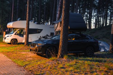 Siyah bir BMW X4 2025. Çimlerin üzerine park etmiş bir çatı çadırı, uzun çam ağaçları ve gün doğumunda ya da günbatımında beyaz bir karavan ile çevrili..