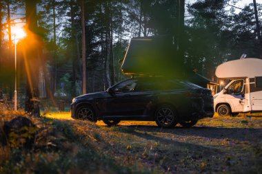 Karanlık bir BMW X4 2025. Ormanlık bir kamp alanında çatı üstü çadırı olan. Sokak lambasından gelen sıcak ışık ve bir karavan uzun çam ağaçlarını aydınlatıyor..