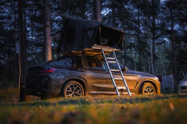 Çatı çadırıyla donatılmış bir BMW X4 2025 günbatımında ormanlık bir alana park edilmiştir. Sahneyi çevreleyen uzun ağaçlar ve arka planda başka bir araç var..