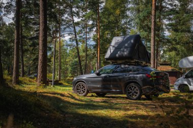 Orman kamp alanında çatısı olan bir BMW X4 2025. Alanı çevreleyen uzun çam ağaçları, dallardan süzülen güneş ışığı....
