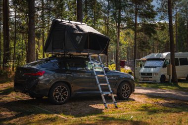 Bir BMW X4 2025. Çatı çadırı ve merdiveni ormanlık bir kamp alanına park edilmiş. Uzun ağaçlar, benekli güneş ışığı ve beyaz bir karavan görülebilir..