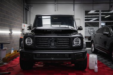 Siyah bir Mercedes G sınıfı SUV, gri tuğlalı duvarlı, parlak ışıklı ve aslan amblemli cam kapılı kırmızı kareli bir garajda park halinde..