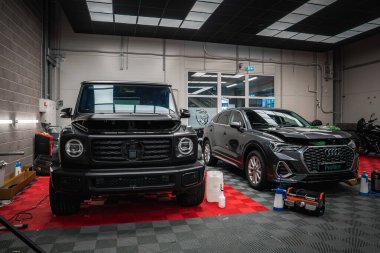 Kaputu açık siyah bir Mercedes G sınıfı ve gri bir Audi SUV temiz, iyi aydınlatılmış bir araba tamirhanesinde kırmızı ve siyah kareli bir zemine park etmiş..