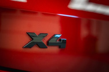 Kırmızı bir BMW 'nin arkasındaki siyah X4 rozetinin detaylı bir görüntüsü. Parlak boya ışığı yansıtıyor, kapalı sergi arka planında bulanıklık var..