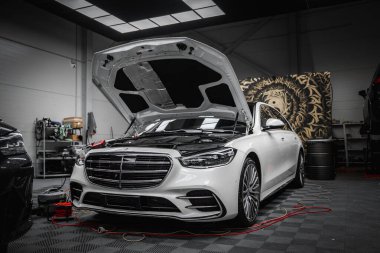 Beyaz bir Mercedes Benz G sınıfı lüks bir araba kaputu açık, temiz, endüstriyel bir garajda kırmızı kablolara bağlı, aletleri ve ekipmanları görünür..