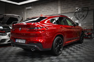 Plaka numarası OD 9766 olan kırmızı bir BMW X4 modern bir garajda kareli bir zemine park edilmiştir. Kaputu açık beyaz bir Mercedes G sınıfı kısmen görülebilir..