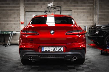 LV OD 9766 plakalı kırmızı bir BMW X4 gri tuğladan duvarları, siyah fayanslı zemini ve duvarında kurt logosu olan şık bir garajda park halinde..