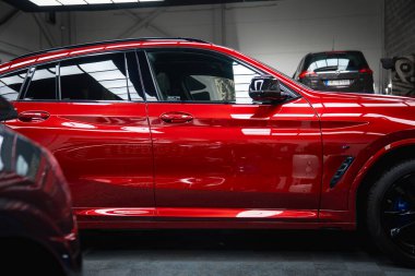 Işıl ışıl bitiren kırmızı bir BMW X4 iyi aydınlatılmış bir garajda park halinde. Siyah Range Rover Discovery endüstriyel iç mekanlarda bir platformda görünür..