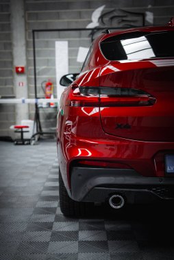 Parlak kırmızı BMW X4 gri tuğladan bir duvarı, yangın söndürücüyü ve arka planda tabureyi olan bir yer. Tepedeki ışıklandırma cilalanmış yüzeyi yansıtır.