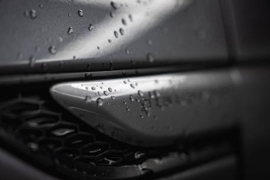 Su damlacıkları, havalandırma ya da ızgara tasarımı ve pürüzsüz metalik hatlara sahip koyu renkli lüks bir araba yüzeyinin detaylı bir görüntüsü..