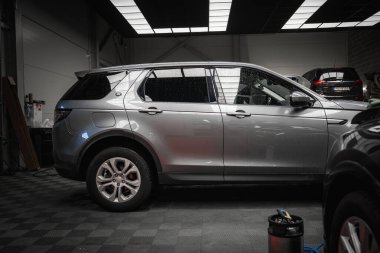 Temiz ve iyi aydınlatılmış bir garajda kareli zemini olan gümüş renkli bir Range Rover Discovery. Araçlar, ekipmanlar ve diğer araçlar kısmen görülebilir.