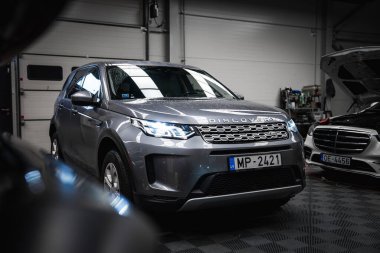 Plakası MP 2421 olan gümüş renkli bir Range Rover Discovery ve temiz, iyi aydınlatılmış bir garajda kaputu açık siyah bir Mercedes G Sınıfı..