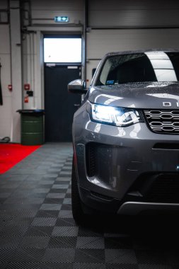 Siyah döşeme üzerine park etmiş farları açık gümüş renkli Range Rover Discovery 'nin önü. Arka planda kırmızı halı ve çıkış işareti var.