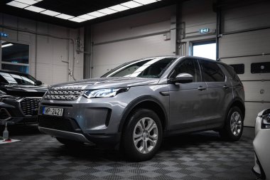 Gümüş renkli bir Land Rover Discovery, fayanslı döşemeleri, parlak ışıklandırması ve BMW X4 ve Mercedes G sınıfı kısmi manzarası olan lüks bir araba garajında park edilmiş..