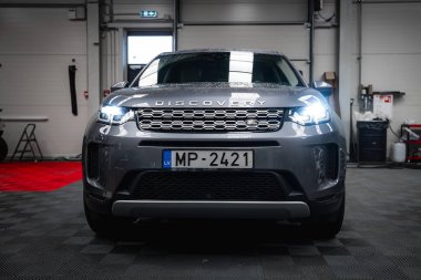 Gri Range Rover Discovery 'nin ön görüntüsü, farları açık, temiz bir garaja park edilmiş, döşenmiş yerler, aletler ve kırmızı halı bölümü var..