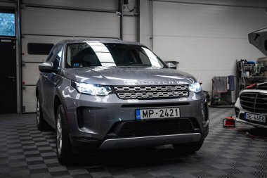 Gümüş Range Rover Discovery, farları açık ve beyaz Mercedes, iyi aydınlatılmış bir garajda kareli zemin ve görülebilir aletlerle kaputu açık..