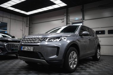 Plakası MP 2421 olan gümüş renkli bir Range Rover Discovery. Kareli zemini, beyaz kapıları ve kısmen görünür siyah Audi 'si olan iyi aydınlatılmış bir garajda..