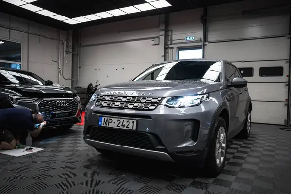 Plakası MP 2421 olan gümüş renkli bir Land Rover Discovery, iyi aydınlatılmış bir garajda. Profilm logosu olan siyah bir Audi sol tarafta kısmen görülebilir..