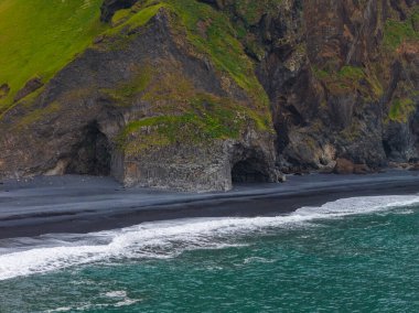 Yeşil kayalıklarla ve bazalt sütunlarla çevrili siyah kumsal. Turkuaz okyanus dalgaları kıyıya vuruyor, çarpıcı bir renk kontrastı yaratıyor.
