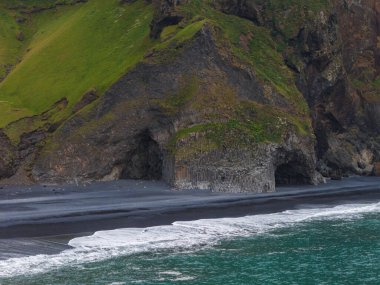 Siyah kumlu bir sahil manzarası, bazalt sütun kayalıkları, yeşil bitki örtüsü ve turkuaz su, İzlanda 'nın volkanik manzarasını gösteriyor..