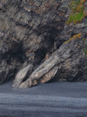 Siyah bir kumsalda katmanlı dokuya ve yeşil bitki örtüsüne sahip pürüzlü kaya oluşumu İzlanda 'nın dramatik kıyı manzarasını gözler önüne seriyor..