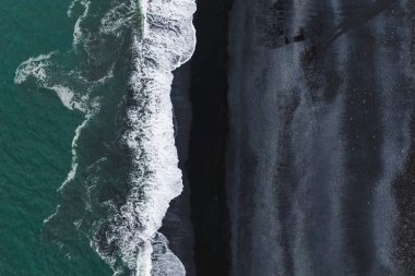 Reynisfjara 'nın İzlanda' daki kara kum plajının hava perspektifinde volkanik kum, turkuaz sular ve kıyı şeridi boyunca beyaz dalgalar yer alıyor..
