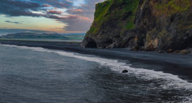 İzlanda 'nın siyah kumsal sahili, yeşil yamalı dramatik uçurumlar, açılış gibi bir mağara, okyanus dalgaları ve günbatımında yumuşak turuncu ve mavi bir gökyüzü barındırır..