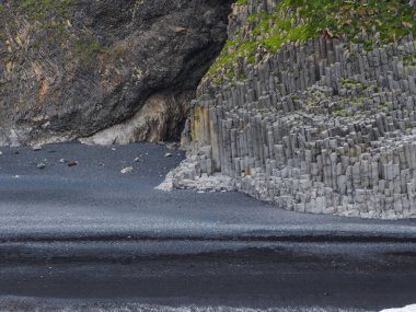 Dikey bazalt sütunlar, engebeli kaya yüzleri ve yeşil bitki örtüsüyle İzlanda kıyı sahnesine doku ekleyerek siyah bir kumsal üzerinde yükseliyor..