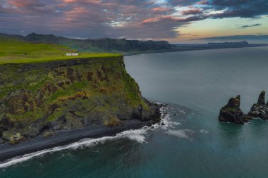 İzlanda 'nın siyah kumsal sahili, sahil kayalıkları bazalt deniz yığınları ve pembe ve turuncu gökyüzünün altındaki yeşil uçurumların tepesinde beyaz bir bina..