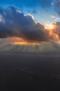 Yeşil bitki örtüsüyle dramatik bir volkanik dağ manzarası, ve bulutların arasından kırılan altın renkleri ile canlı bir günbatımı..