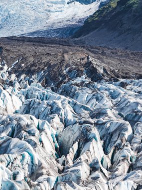 İzlanda 'da derin yarıkları ve mavi buzla koyu moraine zıt bir buzul. Çevredeki dağların yamaçlarında yeşil bitki örtüsü belirir..
