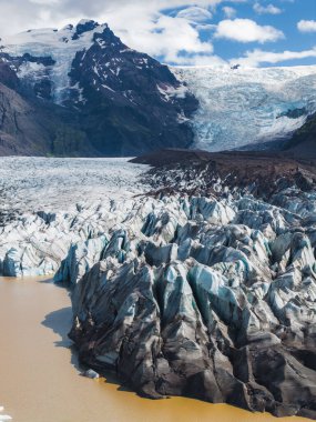 Bir buzulun pürüzlü buz oluşumları, dağılmış bulutlarla kaplı açık gökyüzünün altında, karanlık dağlarla çevrili çamurlu bir buzul gölüyle buluşur..