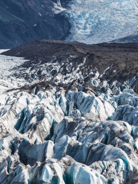 İzlanda 'da derin yarıkları ve koyu volkanik kayalarla çevrili mavi renkleri olan bir buzul. Buzul arka planda geniş bir buz sahasına yayılır..