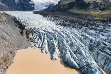 İzlanda 'daki bir buzul, volkanik kaya ve yosun kaplı tepelerle çevrili, arka planda karla kaplı dağlarla kaplı sütlü kahverengi bir erime suyuna dönüşür..