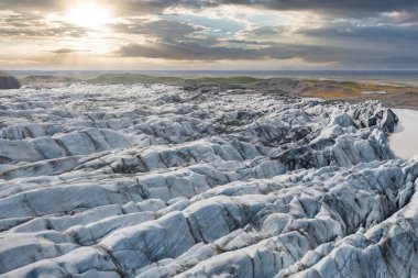 Karmaşık yarıklar, buzlu çıkıntılar, yeşil alanlar ve parçalı bulutlu gökyüzü altındaki engebeli arazi içeren İzlanda 'daki bir buzulun havadan görünüşü..