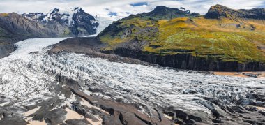 Volkanik arazi ve yosunlu tepelerle çevrili, karlı dağlardan bir buzul akar. Kayalıklar, kayalıklar ve eriyen su havuzları görülebilir..