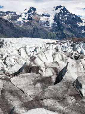İzlanda 'daki bir buzulda, buzlu yüzeyinde koyu volkanik küller, kar kaplı dağlar ve arka planda yeşil karıncalanmış yamaçlar bulunur..