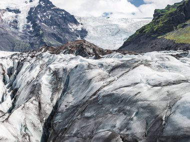 Yeşil yamalı dik volkanik dağlar ve İzlanda 'nın engebeli arazisinde parçalı bulutlu bir gökyüzü tarafından çerçevelenmiş bir buzul ön plana doğru uzanır..