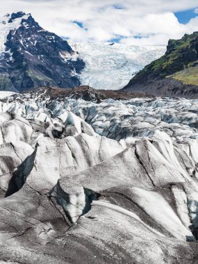 İzlanda 'da koyu volkanik kül çizgileri, arka planda karla kaplı bir dağ ve parçalı bulutlu gökyüzünün altında yeşil yamaçlar olan bir buzul..